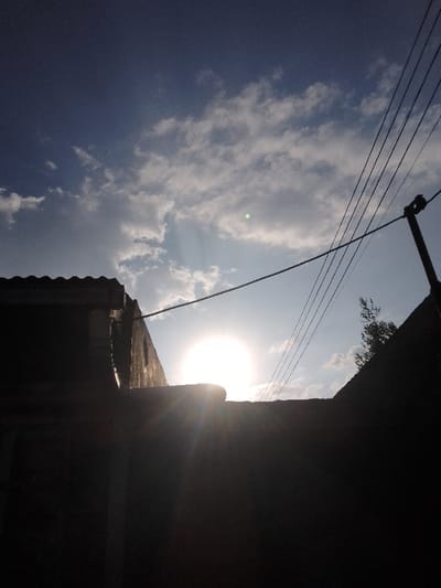 This image depicts an outdoor scene in Njoro ward, Kenya, characterized by strong backlighting from the sun positioned low in the sky, indicating either sunrise or sunset. The upper approximately two-thirds of the frame show a blue sky with scattered white cumulus clouds. The bright, circular sun is prominent in the mid-lower sky, centrally located, generating significant lens flare.
The lower portion of the image is dominated by dark, silhouetted structures. A solid, dark, horizontal structure forms the immediate foreground. To the left, a building's silhouette is visible, featuring a corrugated or tiled roofline and a vertical pipe or downspout on its side. To the right, another dark, angled structure, possibly a building or wall, is present. A dark utility pole stands on the far right, extending vertically from this angled structure, supporting multiple parallel power lines that stretch diagonally across the mid-sky towards the center-left. A small cluster of dark foliage, consistent with tree leaves, is visible on the far right, above the angled structure. No individuals or discernible activities are present.