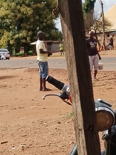A group of people are gathered in a dirt-filled area next to a road. A man in a yellow shirt and blue shorts stands on the left, facing right. To his right, another man in a dark t-shirt and white shorts is visible, holding a white bucket. A third person in a green shirt is further in the background, walking away from the viewer. In the background, a white car is parked on the side of the road, and a building with a thatched roof is visible. A motorcycle is parked in the foreground on the right side of the image. The ground is a mix of dry soil and small stones. The environment appears to be rural or semi-urban, likely in Ichol, Nigeria, given the context.