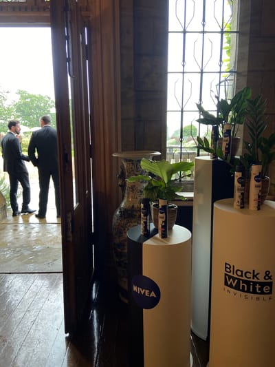 An indoor view looks out through an open wooden doorway and a window with a decorative metal grate. Two men in dark suits stand on a wet stone patio outside, their backs to the viewer, with trees and white umbrellas visible in the background. Inside, to the right, are several display stands featuring Nivea Men Black & White Invisible deodorant spray cans. One stand is tall and white with the "Black & White Invisible" text prominently displayed. Another stand, partially visible, is a shorter, two-toned cylinder with a Nivea logo. Potted plants are placed on and around the display stands. A large, ornate vase is positioned to the left of the display. The floor inside appears to be dark wood and is also wet. The overall impression is of an event or product display, possibly at a venue in Chichester, United Kingdom, given the architectural style and exterior setting.