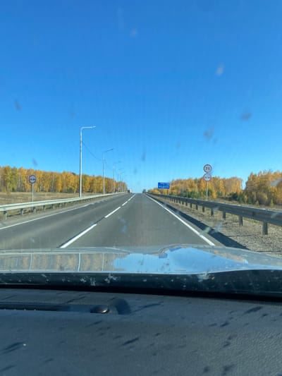 This image captures a multi-lane highway near Kuytun, Russia, on a clear autumn day, viewed from the interior of a vehicle. The foreground shows the dark-colored, textured dashboard and a reflective chrome-like strip, which mirrors the blue sky and distant foliage. The windshield, through which the scene is observed, exhibits several circular dust or smudge marks.
The asphalt highway stretches directly ahead, presenting two visible lanes marked by dashed white lines, bordered by a solid white line on the right shoulder. Metal guardrails line both sides of the road. On the left, a series of evenly spaced streetlights with slim poles recedes into the distance. A circular white sign with a red border on the left side depicts two red vehicle silhouettes—a passenger car and a lorry—positioned side-by-side within the circle. Beyond the guardrail, a dense stand of trees displays vivid yellow and orange autumn foliage.
On the right side of the road, two stacked circular signs are present. The upper sign is white with a red border and displays "70" in black, indicating a 70 km/h speed limit. The lower sign is a blue circle with a red border and a single red diagonal line, signifying "No Stopping" (Russian sign 3.27). Further along the highway, a large blue rectangular informational sign with white text is mounted on an overhead structure. A similar line of bright autumn trees borders the right side of the road.
In the far distance, several vehicles, including what appears to be a large truck, are visible on an ascending section of the highway. The sky is uniformly brilliant blue and entirely cloudless, indicating direct sunlight conditions.