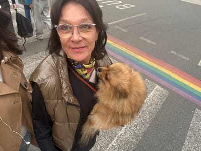 An eye-level outdoor image captures a middle-aged woman, a dog, and a partial view of another person on a street in Blois, France. The central subject is a woman with dark hair, wearing silver-rimmed glasses with a slight yellow tint, an olive-green puffer vest over a dark long-sleeved top, and a yellow, multi-colored patterned scarf. She exhibits a faint smile while looking directly at the camera. Nestled in her arms or a carrier is a fluffy, light brown Pomeranian dog, its head turned to the right, away from the viewer. To the immediate left, a portion of another individual is visible, wearing a light brown trench coat, with white wired earphones visible near their chest. In the blurred background, several figures are present, including one carrying a black backpack. The ground consists of dark asphalt featuring a prominent rainbow-colored pedestrian crossing, composed of horizontal stripes (red, orange, yellow, green, light blue, purple). White parallel pedestrian crossing lines and dashed road markings are also visible. The partial word "BUS" is stenciled in white on the asphalt in the upper right quadrant of the image.