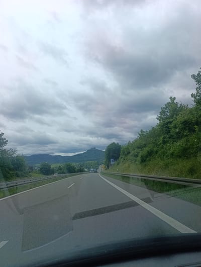 The image presents a view from inside a vehicle traveling on a multi-lane road, likely a highway, under an overcast sky. The dark asphalt road, marked with a solid white line on the right and a dashed white line for the center divider, extends into the distance. Reflections are visible on the vehicle's windshield in the foreground. A metal guardrail runs along the right side of the road, bordered by dense green trees and bushes that ascend a gentle slope.
In the midground, the landscape features rolling hills and mountains, all covered in abundant green foliage. Perched atop a distinct, prominent conical hill in the distance, a large, dark, castle-like structure with multiple turrets and spires is visible, a notable architectural landmark.
The sky is heavily covered with a blanket of grey and white clouds, indicating an overcast or cloudy day with diffuse lighting. On the right side of the road, a blue rectangular sign with a white 'P' symbol (indicating parking) and the text "300m" is clearly visible, suggesting an upcoming parking area. No people are explicitly visible in the scene, and no specific time of day beyond daytime is discernible due to the cloud cover. The overall setting appears to be a scenic, hilly region, consistent with parts of Germany.