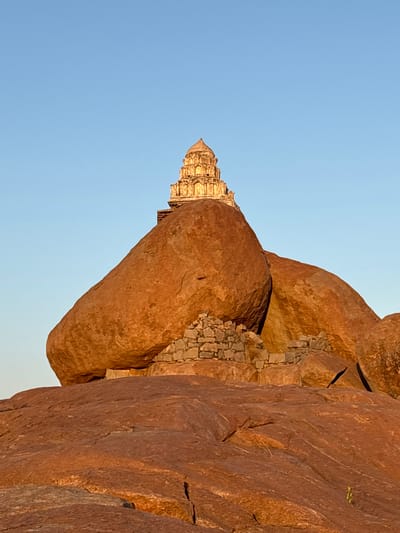 The image shows the Achyutaraya Temple, a ruined temple complex in Hampi, near Kamalapura, India. The temple is partially obscured by large, reddish-brown boulders typical of the Hampi landscape. A small portion of the blue sky is visible in the upper left corner of the image. The temple itself is a light color and intricately carved. Smaller stones appear to be wedged between the boulders.