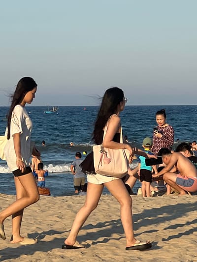 The image shows a group of people on a sandy beach with the ocean in the background. The sun appears to be low in the sky, casting long shadows, suggesting it is either early morning or late afternoon. The water is a deep blue, with waves gently rolling onto the shore. Several individuals are visible, some walking along the sand, others interacting near the water's edge. One woman in the foreground is facing away from the camera, looking towards the ocean, while another woman is closer to the background, also looking out at the sea. Some individuals are wearing swimwear, while others have casual clothing on. The overall atmosphere seems relaxed and summery, with people enjoying a day at the beach. There are no visible text elements in the image. The location context provided is Đà Nẵng, Vietnam.