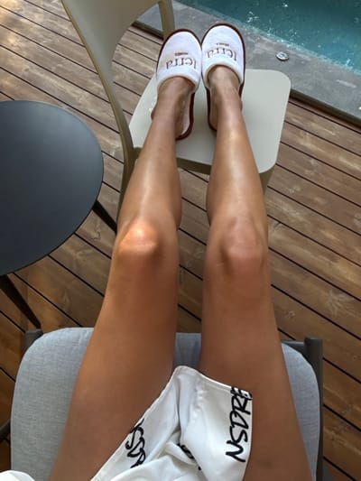 This is a first-person perspective image of a person's legs and feet, wearing white slippers with brown trim. The slippers have the word "terra" and "HOMES" printed on them in a brown font. The person is seated on a gray chair on a wooden deck. To the left of the frame is a dark gray circular table. In the background, a swimming pool with blue water is visible. The scene is set in Bakovići, Montenegro.