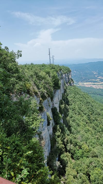 The image captures a high-angle, exterior view of a steep, rocky cliff face covered in lush green vegetation. The cliff, composed of layered gray rock, has patches of greenery growing from its crevices. At the top of the cliff, several tall, metallic telecommunication towers are visible against a bright, hazy blue sky with scattered white clouds. The surrounding landscape is densely forested with trees, and in the distance, a valley opens up, revealing a small town with red-tiled roofs and agricultural fields, indicating the location is near Susqueda, Spain. The image does not contain any people or discernible actions.