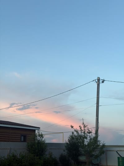 A photograph captures a suburban scene in Nizhny Suyk-Su, Russia, under a pastel-colored sky at dusk. The sky is a gradient of light blue at the top, transitioning to soft pink and pale orange hues near the horizon, with wispy clouds scattered throughout.

In the foreground, a section of a wooden house with a dark brown, horizontally-paneled exterior is visible on the left. It has a dark-framed window and a white trim along the roofline. A grey metal corrugated fence runs horizontally across the lower part of the image, separating the property.

To the right of the house, a wooden utility pole stands tall, supporting multiple black electrical wires that stretch across the frame. A small, leafy green tree is situated in front of the pole and to the right of the fence. Further to the right, a large green barrel is partially visible at the bottom edge of the frame. The overall impression is one of a quiet, rural or semi-rural evening setting.