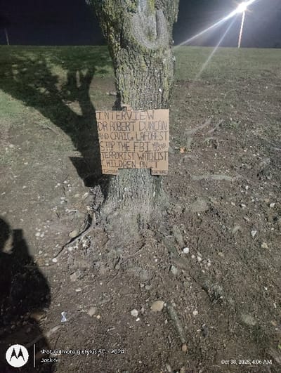 The image displays an outdoor, nighttime scene centered on a large tree trunk with a handwritten cardboard sign affixed to it. The tree's bark is textured and appears to have moss or lichen. The light brown, rectangular sign, oriented vertically, is secured to the tree and features text written in a dark marker. The legible text on the sign states: "INTERVIEW DR ROBERT DUNCAN AND CRAIG LAFOREST STOP THE FBI MK ULTRA TERRORIST WATCHLIST CHILDREN ON IT." The word "TORTURE" is printed in smaller letters at the bottom left and right corners of the sign.

The ground surrounding the tree consists of dark, disturbed soil mixed with exposed roots, small rocks, and sparse debris. To the right of the tree, a gently sloping grassy area is visible. A bright, artificial light source, characteristic of a streetlamp, is prominent in the upper right quadrant, casting strong rays and illuminating parts of the scene. In the lower left foreground, a distinct shadow of a person holding a mobile device is cast, indicating the photographer's position. The image metadata shows a capture date of October 30, 2025, at 4:06 AM. The precise location within the city of Tulsa, United States, cannot be determined from the visual information provided.