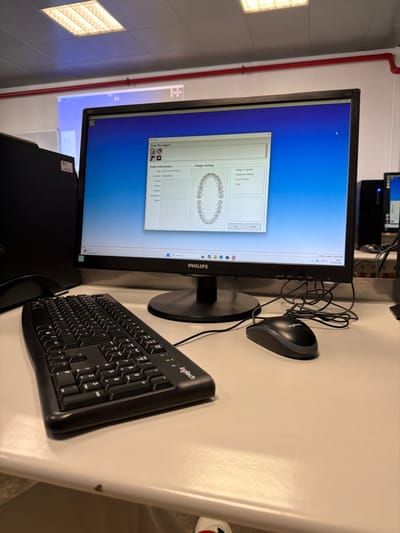 The image displays a computer workstation located in Gandra, Portugal. A black Philips monitor prominently shows a "Scan File Import" software interface with "Order Information" fields, including a date and time of "2020-02-04 09:56:25" and an "Order ID: 29030040586". The "Design Setting" section features a detailed diagram of upper and lower human dental arches, indicating individual teeth and small circles, possibly representing missing teeth or potential implant locations. Options such as "Bridge / Copings" and "Diagnostic Waxup" are visible. A black desktop computer tower is positioned to the left of the monitor. On the light-colored desk surface in front of the monitor are a black Logitech keyboard and a black Logitech wired mouse. Overhead, rectangular fluorescent light fixtures are visible, and a red pipe runs horizontally along a white wall in the background. A blurry projection screen or white board is partially visible behind the monitor. To the right, another computer setup is partially visible. A portion of a person's foot, wearing a white shoe with a red accent, is visible at the very bottom of the frame.