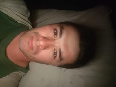 A man lies on a pillow, gazing into the camera. He wears a green shirt and light-colored sweater. His dark hair is short. His facial features, including his nose, lips, and eyes, are clearly visible, and his skin has a reddish tone. The lighting is dim, casting shadows on his face and the pillow.