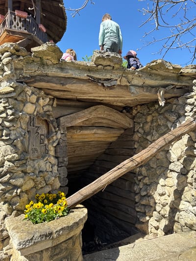 This eye-level, vertically-oriented photograph shows a well built of stone and wood at the Ethno Complex in Kalimantsi, Bulgaria. The well's opening is dark and enclosed by stone. A wooden beam supports part of the stone structure. Yellow flowers grow from a stone fixture at the well's edge. "1974" is carved into a stone embedded in the well's lower section.

At the top of the well, partially visible from the viewer's perspective, are three people: a child with pink hair and a woman. A man wearing a green shirt is seen on the top right-hand edge. The sky, along with some tree branches, is visible in the upper left-hand corner. Another building is visible in the bottom-left corner.