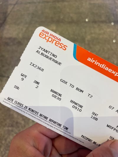 The image shows a close-up of an Air India Express boarding pass. The pass is primarily white with orange and blue accents for the airline's logo and branding. The text on the pass includes the passenger's name (JYANTINO ALBUQUERQUE), flight number (IX2368), origin and destination (GOX to BOM), terminal (T2), boarding time (0830), departure time (0910), seat number (18B), and gate number (9). Additional information includes a passport number and a statement regarding gate closure and baggage checking procedures. The phrase "GATE CLOSES 25 MINUTES BEFORE DEPARTURE TIME" is prominently displayed. The boarding pass is being held by a person.