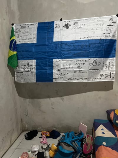 An interior room in Desa Batu Layar, Indonesia, is depicted, showing a bare, light gray concrete wall with a visible corner. A large Finnish flag, predominantly blue and white, is stretched horizontally across the upper wall, secured by black clips at its top corners. This flag is extensively covered with handwritten messages, names, and simple drawings in black marker. Partially overlapping the Finnish flag on the upper left is a smaller segment of a green, yellow, and blue flag, consistent with the flag of Brazil. The lower section of the wall exhibits minor cracks and discoloration. The floor is covered with light-colored tiles. In the foreground, a collection of personal items is arranged on the floor: black clothing items, various snack packets, folded fabrics, a clear plastic bag containing white objects, several bottles, a light brown plush toy, a blue and gray backpack, and a pair of pink and gray athletic shoes. A small pink rectangular object leans against the wall behind the backpack. To the far right, a bed or mattress with a brightly patterned cover featuring geometric shapes in blue, orange, and white is partially visible. No individuals are present.