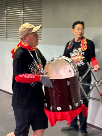 Two men are holding a drum. The man in the foreground is wearing a beige baseball cap, black t-shirt with colorful design, gray gloves, red arm sleeve with Chinese characters, and a red scarf. He is holding the drum with both hands. The other man is wearing a black t-shirt with colorful design and a red arm sleeve. He is standing behind the drum, holding a pair of drumsticks. A red cloth is wrapped around the drum. The location is Longxing, China.