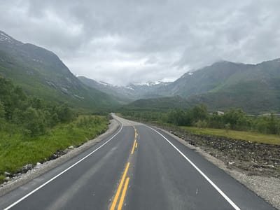 The image depicts a paved road curving through a mountainous landscape under an overcast sky. The road has white lines marking the edges and yellow double lines indicating the center. To the left of the road, a grassy slope rises, dotted with trees and leading to a steep, green mountainside. The right side of the road features a rougher embankment of soil and rocks, with sparse vegetation. In the distance, the road leads towards a valley framed by more mountains, some of which have patches of snow on their peaks. The overall scene suggests a remote, natural setting, characteristic of the region around Gullesfjord, Norway. There are no people or vehicles visible in the image.