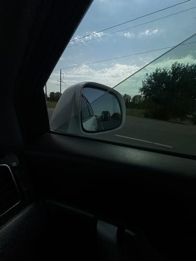 The image is a view from inside a car, looking out the passenger window towards the side mirror and the road. The side mirror reflects a truck driving on a road. The car appears to be in motion, as indicated by the motion blur in the reflection and the scenery outside. The environment suggests a semi-urban or rural setting, with trees and power lines visible along the roadside. The sky is partly cloudy, with the sun partially obscured. There are no people clearly visible in the image, only the interior of the car and the exterior scene. The image was taken in Istiqlol, Uzbekistan.