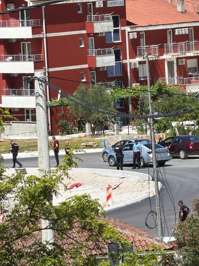 The image depicts a bright, sunny daytime street scene, likely in Kubasi, Montenegro, featuring a notable police presence. In the center-right, two police officers stand near a silver sedan with its front driver's side door open. One officer wears a light blue shirt and dark pants, while the other is in a darker uniform. A third individual, also in dark attire, is further back near the silver car. Behind it, a dark red sedan is parked.
On the left side of the frame, a prominent multi-story building with reddish-brown walls and white trim features numerous balconies with white railings, windows, and external air conditioning units. The text "APARTMENTS GOLD" is clearly visible on its side. The building is topped with a terracotta-tiled roof.
Several other individuals are present on the pavement and street. One person in a red shirt and dark pants stands on the pavement, while another in a dark t-shirt and cap is further down the road. A utility pole with numerous cables runs vertically through the scene. A red and white striped barrier lies on the ground near the silver car. Green trees and bushes, some with pink flowers, are scattered throughout, and strong sunlight casts clear shadows, indicating good weather. The scene suggests an official incident or police activity on a residential or commercial street.