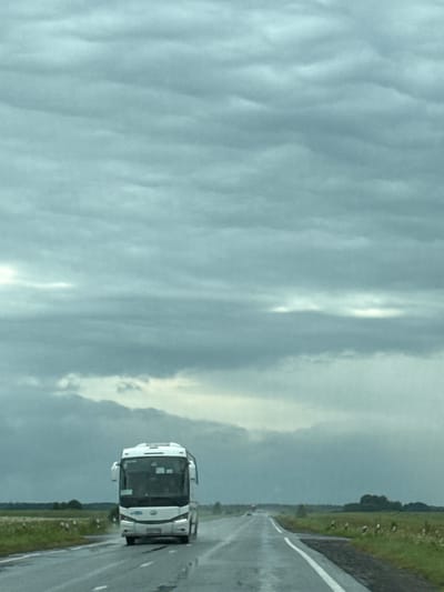 A white tour bus is driving on a wet asphalt road in Deevo, Russia. The sky is cloudy and the weather appears to be stormy. The road is flanked by grassy fields and trees. The bus is the main subject of the image and is positioned in the center of the frame, heading towards the viewer. The scene is set in a rural or suburban area.