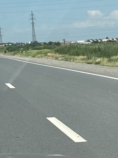 The image shows a straight asphalt road with white lane markings. The road curves slightly to the right from the perspective of the viewer. In the background, there is a grassy embankment with some low vegetation and a few structures that appear to be rural buildings or industrial facilities. A tall, lattice-like electrical pylon stands prominently in the middle ground. Overhead, multiple power lines stretch across the frame. The sky is blue with scattered clouds. The scene is devoid of any people or vehicles. The location is Taperakan, Armenia.