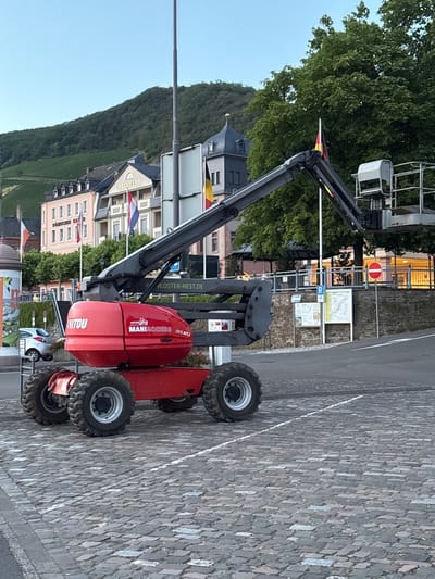 In Bernkastel-Kues, Germany, a red Manitouski 120 ATJ aerial work platform is parked on a cobblestone surface. The machine is facing right, with its boom extended upwards and to the right, holding a work platform. In the background, a row of historic buildings with pitched roofs and flags lining the street are visible. A lush green hillside with vineyards rises behind the buildings. The sky is clear and blue. The scene appears to be a public area, possibly a town square or waterfront, given the cobblestone paving and the presence of buildings and a road.