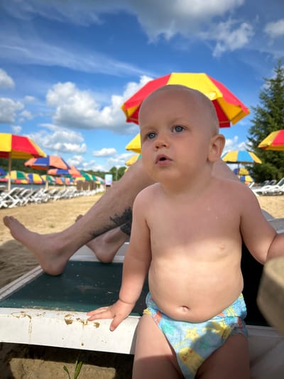 A close-up, low-angle shot shows a young child with blue eyes and fair skin, wearing swim trunks with a pattern of yellow starfish and other sea creatures. The child's mouth is slightly open, as if speaking or surprised. Part of an adult's legs, with visible tattoos on the shin, are also in the frame, suggesting they are holding or sitting next to the child.

The background is a bright blue sky with fluffy white clouds. Several colorful beach umbrellas with alternating red and yellow panels are visible, indicating a sunny outdoor setting. Some of these umbrellas are clustered together, and others are spread out further down what appears to be a sandy beach. In the distance, more umbrellas and some foliage can be seen. The child is positioned on what looks like the edge of a lounger or a similar piece of beach furniture.

The overall impression is one of a summer day at a beach resort, possibly in Юбилейный, Russia, given the location context. The lighting suggests it is daytime, likely late morning or early afternoon due to the bright sunlight casting shadows. The child's expression and the relaxed pose of the adult's legs convey a sense of leisure and enjoyment. No visible text is present in the image.