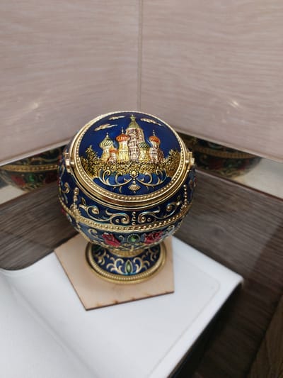 A darkly ornate, spherical object, possibly an ashtray or decorative box, rests on a white surface in a corner. The object is predominantly dark blue with intricate gold embellishments and features a detailed depiction of a multi-domed building, likely St. Basil's Cathedral in Moscow. The background consists of light-colored tiled walls and dark wood paneling. No people or actions are depicted, and the image appears to be a still life. There is no information to indicate the location in the city of Nakhabino, Russia.