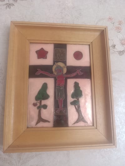 A framed artwork depicting a stylized crucifixion is displayed against a floral-patterned wallpaper. The artwork is centrally positioned and framed by a light-colored wooden frame. The central image features a figure on a cross with outstretched arms. Above the figure's head, the words "JESUS" and "REX MUNDI" are inscribed. The artwork is divided into quadrants, with symbolic elements like a star and a circle present in the upper sections, and trees in the lower sections. The colors within the artwork are rich, with reds, greens, and metallic tones. The wallpaper behind the artwork features a subtle pattern of leaves and flowers in muted pink and white tones. The overall scene is static and the artwork appears to be a decorative piece. The image does not provide any information about the city of Berching, Germany.