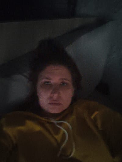 A woman with medium-length brown hair is lying down, looking directly at the camera. She has blue eyes and is wearing a yellow hooded sweatshirt. The lighting is dim, casting shadows on her face and the background. The image has a grainy texture, suggesting it may have been taken in low light conditions or with a camera that produces significant noise. The background appears to be a dark, indistinct space, possibly an interior. There is no visible text in the image. The overall mood is somber, potentially due to the lighting and the woman's neutral expression. The context of Kohtla-Järve linn, Estonia, does not provide specific visual cues within the image itself.