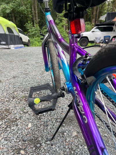An extreme close-up shot captures the rear of a purple and teal ombre bicycle parked on a gravel surface, likely in a campsite in Chilliwack, Canada. A portion of the bicycle's black pedal with a yellow circular sticker is visible in the foreground. In the background, out of focus, a white SUV is parked, and a section of a green and gray tent is visible. The environment appears to be a wooded area with trees in the background.