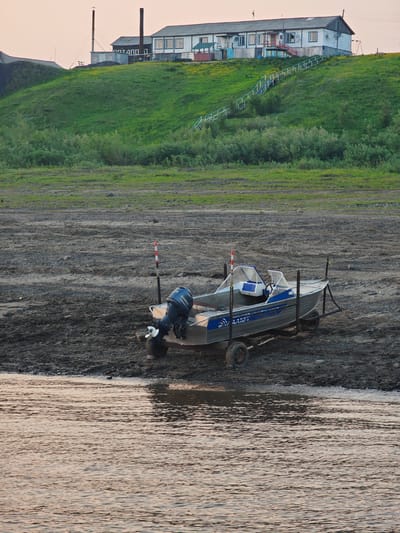 This is a forensic, matter-of-fact description of the provided image, which depicts a scene in Potapovo, Russia.
The image showcases a small, aluminum motorboat resting on a trailer on a gravelly shore. The boat is a light silver color with blue accents, featuring a dark blue outboard motor labeled "YAMAHA" on the stern. The interior of the boat has blue seating and a steering console. The trailer has two wheels on each side and is positioned on the shore, which is composed of mud, gravel, and scattered rocks.
Behind the boat, the shoreline extends towards a grassy hill. The hill is covered in green vegetation and dotted with small yellow flowers. On top of the hill sits a long, white, multi-story building with a gray roof. The building has multiple windows and doors, and a red-railed staircase leads up the hill to the building's entrance. Adjacent to the building are two tall, dark chimneys, suggesting industrial or heating facilities.
The foreground of the image shows a body of water with ripples reflecting the soft, possibly evening, light. The water appears calm, with subtle golden and pink hues. The environment suggests a rural or semi-industrial setting near a body of water, consistent with a location in Potapovo, Russia. No people are visible in the image.