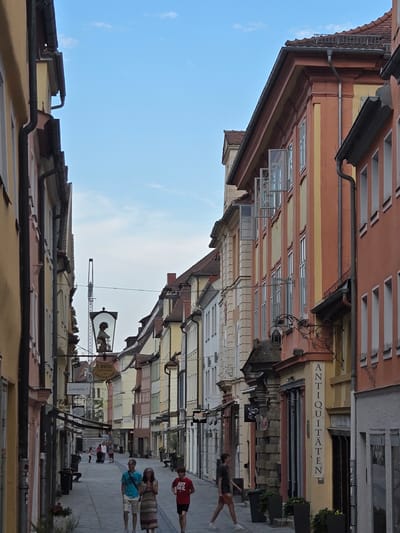 This image depicts a street scene in Ansbach, Germany, characterized by narrow buildings with colorful facades and an overcast sky. Several people are visible walking along the cobblestone street. One person in a red shirt with "JUST DO IT" printed on it is walking away from the camera. Another person wearing a blue shirt and shorts is walking towards the camera, accompanied by two other individuals. In the background, more people are scattered along the street.
The architecture features traditional European styles with multiple stories, numerous windows, and decorative elements. Signs are visible on the buildings, though some are partially obscured or difficult to read. One sign appears to read "ANTIQUITÄTEN", indicating an antique shop. Another sign is a circular illuminated sign with a silhouette of a person. The overall impression is of a lively pedestrian area within a historic town. The lighting suggests it could be daytime, possibly late afternoon or a cloudy day given the lack of harsh shadows. The atmosphere seems relaxed and ordinary, with people going about their day.