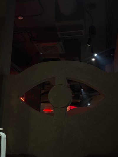 The image shows an interior scene with a dark, moody ambiance, likely a restaurant or bar. In the foreground, a large, curved concrete structure with an eye-shaped cutout dominates the frame. Through the cutout, glimpses of red lighting and what appear to be ceiling fixtures are visible. The ceiling above is partially visible, showing electrical conduits, a smoke detector, and HVAC units. A few track lights are also present, casting bright spotlights. The overall impression is one of modern, industrial-inspired decor with dramatic lighting. The location in Borodino, Russia, is not visually discernible from the image itself, as there are no specific architectural features or signage that would indicate this location.