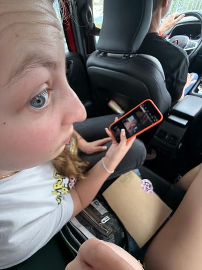 A person is taking a selfie while sitting in a car. The person is holding a cell phone in their right hand, looking at the screen. Another person is driving the car. There is a purse and an envelope on the passenger seat. The location is indeterminate, but the city is Chokoba, Bulgaria.