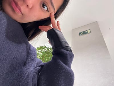 A selfie looking upward features a person with long, dark hair and fair skin wearing a dark blue sweater. Their finger touches their eyebrow. The background reveals a low-angle view of a white wall with a green and white emergency exit sign. Above the wall, green foliage is visible, possibly from trees. The unusual perspective suggests the photo was taken from below, pointing upwards.