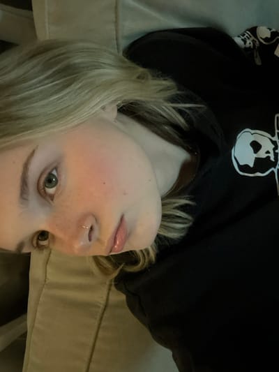 A pale-skinned person with light blonde hair and green eyes is looking at the camera. They have freckles and a lip ring. They are wearing a black hooded sweatshirt with a white graphic on the shoulder. The background is a neutral-colored, possibly beige, cushioned surface. The image is rotated 180 degrees, presenting an upside-down perspective. The lighting is soft and diffused.