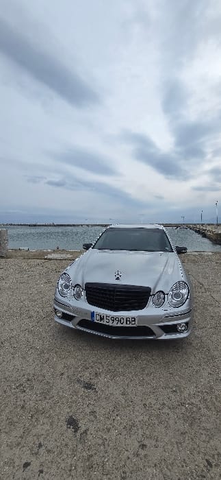 A silver Mercedes-Benz E-Class sedan, W211 generation, is parked facing the viewer on a textured concrete and gravel surface in Limenas Avdiron, Greece. The vehicle displays a Bulgarian license plate, "CM 5990 BB." It features a black grille with a prominent central Mercedes-Benz star emblem, projector headlights, and an aftermarket lower front bumper with integrated circular fog lights. A dark tinted strip is applied to the upper portion of the windshield.
Behind the vehicle, a calm body of water, consistent with a sea or harbor, extends towards the horizon. The shoreline consists of concrete and scattered gravel. To the right, a stone or concrete breakwater or pier extends into the water. A large, light-colored concrete pillar stands to the left of the car. The sky is heavily overcast with multiple layers of grey clouds, indicating muted daylight. No individuals are visible in the scene. The concrete ground beneath the vehicle exhibits minor dark stains.