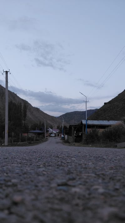 The image captures a low-angle view of a paved road leading into a village in Ak-Terek, Kyrgyzstan. The road surface in the foreground is rough and textured with visible stones. On either side of the road, rolling hills covered in sparse vegetation rise upwards. The sky is overcast with subtle cloud formations, suggesting either early morning or late evening.

Along the road, on the left side, a tall utility pole stands with multiple wires extending outwards. Further down the road, houses with dark roofs and fences are visible, interspersed with trees. A white car is parked on the road. On the right side of the road, there are more structures, including a building with a corrugated metal roof supported by blue posts, and dense bushes. The overall scene depicts a quiet, rural setting within a mountainous region. There are no people visible in the image.