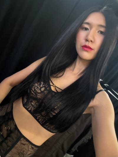 A single young adult female is depicted in a medium-shot selfie, facing forward, holding a camera or mobile device at arm's length. She has long, straight black hair extending over her shoulders and chest. Her lips are covered in bright pink lipstick. She is wearing a black two-piece lace lingerie set; the top is a bralette with intricate floral patterns, beige lining, thin straps, and a small gold-colored ring detail at the décolletage. The bottom is a high-waisted lace piece with similar floral patterns and small gold-colored rings along what appears to be a waistband or garter attachment. The background consists primarily of dark, vertically draped fabric, likely a curtain or backdrop, suggesting an indoor setting. A dark, reflective surface or object is partially visible on the right side of the frame. The specific location within Herentals, Belgium, cannot be determined from the provided image.