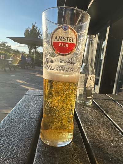 This image depicts an outdoor patio setting, characteristic of a public house or restaurant garden, likely located in Brandesburton, United Kingdom.
In the foreground, a tall, clear glass, approximately three-quarters full of effervescent golden liquid consistent with lager, stands upright on a dark, textured outdoor table surface. The glass features a red and gold "AMSTEL BIER" label and "1870" embossed near its base. To the right of the glass, an empty, clear glass Corona Extra beer bottle (33cl/330ml) is also standing on the table.
The midground to the left reveals a light-colored paved patio area illuminated by natural sunlight, causing lens flare. Several indistinct, blurred figures are seated at dark outdoor tables under a large, dark patio umbrella, suggesting social interaction. To the right, a dark building structure, possibly the exterior of the establishment, is visible. Beyond the patio and seated figures, trees and foliage are present against a clear blue sky with faint clouds, indicative of a late afternoon or early evening setting due to the low angle of the sun.