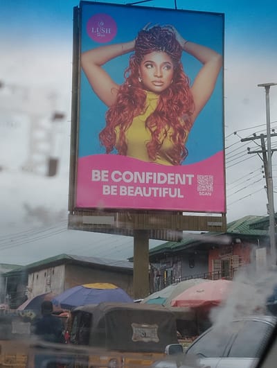 A medium shot captures a billboard displaying a woman with elaborate red hair and braids, posing with her hands on her head against a bright blue sky. The billboard features the logo "LUSH Hair" in a pink circle and the text "BE CONFIDENT BE BEAUTIFUL" in white. A QR code with the word "SCAN" is also visible on the pink section of the billboard. The billboard is supported by a metal structure.
Below the billboard, the scene depicts a street in Aketu-Oja, Nigeria. Several market stalls with colorful umbrellas are set up, including blue, yellow, and red ones. A person is seen standing near some of the stalls. Various vehicles, including what appears to be an auto-rickshaw and a car, are present on the street. Buildings with green roofs and signs of commercial activity line the background. The overall impression is a bustling street scene with a prominent advertising billboard.