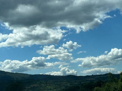 The image depicts a landscape scene with a dense, dark green, tree-covered mountainous terrain in the foreground. Above the terrain, the sky is a vibrant blue, populated with numerous cumulus clouds of varying sizes and shades. Some clouds are bright white and fluffy, indicative of fair weather, while others are darker and more ominous, suggesting potential rain or stormy conditions. The overall impression is that of a scenic, natural environment, possibly in a rural or mountainous region, under a dynamic sky. The location is identified as Novachene, Bulgaria.
