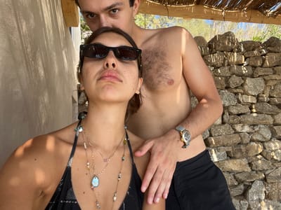 The image depicts two individuals, a female and a male, in a sunlit outdoor setting, identified as Agios Ioannis Diakoftis, Greece. The female is positioned in the foreground, facing towards the viewer, wearing dark rectangular sunglasses, a black bikini top with white patterns, and multiple layered necklaces including a prominent light blue oval pendant. Her expression is neutral, and her head is tilted slightly upwards. The male stands directly behind her, bare-chested with visible chest hair and wearing dark swim shorts. His right hand rests on the female's left hip/lower back. He wears a gold-toned wristwatch with a light strap on his left wrist. His face is visible above the female's head, looking directly towards the camera with an intense expression.
The background features a light-colored, possibly stucco or concrete, wall on the left, displaying distinct shadows from an overhead structure. On the right, a dry-stacked stone wall is visible. Above the individuals, a thatched roof supported by wooden beams provides shade, characteristic of a pergola or cabana. Beyond the stone wall, distant vegetation and a clear sky are discernible. The scene is illuminated by bright natural light, casting sharp shadows, indicative of daytime.
