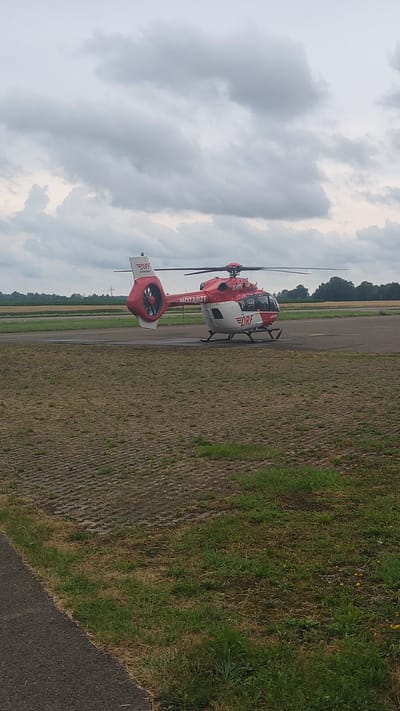 The image shows a red and white helicopter with the text "DRF" and "NOTARZT" visible on its side. The helicopter appears to be a medical helicopter, likely an air ambulance, given the "NOTARZT" (German for "emergency doctor") text. It is parked on an asphalt surface, possibly a helipad or an airfield, with a grassy area and a field visible in the background. Trees and a cloudy sky suggest an outdoor setting. The overall scene implies a moment of readiness or standby for the helicopter, perhaps waiting for a call or having just completed a mission. The lighting and cloudy sky suggest it is daytime, and the presence of green grass indicates a temperate climate. The location context provided is Genderkingen, Germany, which helps to place the scene. There are no people clearly visible in the image.