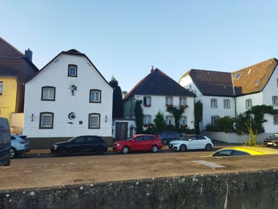 The image shows a row of white buildings with tiled roofs, characteristic of older European architecture, lining a street in Leverkusen, Germany. The buildings have numerous windows and some are adorned with ivy and flower boxes, suggesting a residential area. Several cars are parked along the street. From left to right, a dark gray van, a light blue SUV, a black hatchback, a red station wagon, a dark gray sedan, a white hatchback, and a dark brown sedan are visible. The sky is clear and blue, indicating daytime. The street appears to be paved, and there is a concrete wall in the foreground, suggesting a slight elevation change. The overall scene is peaceful and orderly.