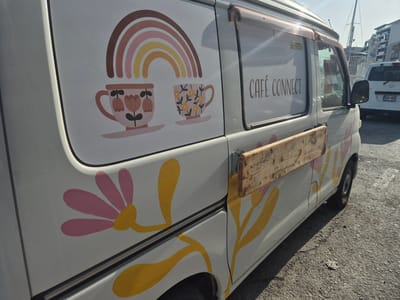 The image shows the side of a white food truck parked on an asphalt surface. The truck has decorative elements including a rainbow and two teacups with flowers. The text "CAFÉ CONNECT" is written on the side of the truck. There is a wooden plank attached to the side of the truck. In the background, a white vehicle with license plate "OGV 669" is visible, along with some boats and buildings. The scene appears to be in Ta' Xbiex, Malta, based on the context provided. The lighting suggests it is daytime.