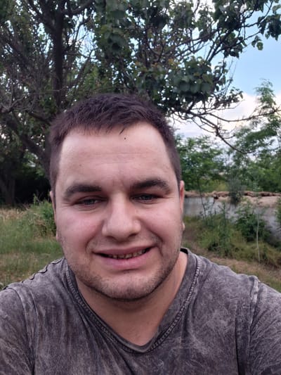 A man is looking at the camera, smiling. He has short brown hair and blue eyes. He is wearing a gray t-shirt. The background is out of focus and shows trees and foliage. The image was taken outdoors.
