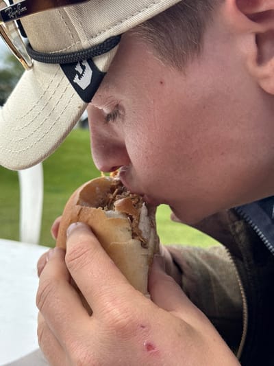 A young person, wearing a white baseball cap with a black "U" logo, is biting into a sandwich with both hands. They are seated at a table outside. The sandwich appears to be filled with pulled pork. The person is wearing a navy blue and green jacket,  and a white baseball cap with brown sunglasses on top of it.  The background appears to be a green field.