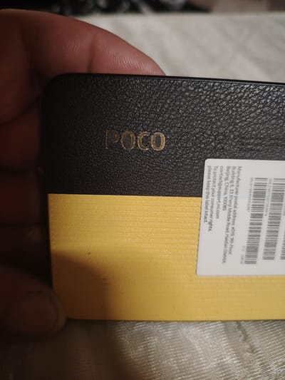 A close-up shot shows a person's thumb and index finger holding the corner of a black and yellow rectangular object. The object has "POCO" embossed in gold lettering on the black portion, and a white label with text and a barcode is affixed to the lower right corner of the black section, overlapping onto the yellow section. The yellow section has a ribbed texture. The background is blurred and appears to be a textured surface, possibly a blanket or fabric. The lighting suggests indoor conditions. There is no visible connection to the city of Gorshki, Russia.