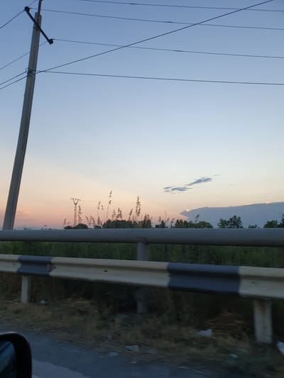 The image captures a view from a moving vehicle on a road in Apaga, Armenia. The scene depicts a rural landscape at dusk. A utility pole with overhead wires is prominent on the left side of the frame, stretching diagonally upwards. In the distance, a power transmission tower with wires is visible, silhouetted against the soft pastel colors of the sunset sky, which transitions from pale orange and pink near the horizon to a clear blue higher up. A reflective guardrail with black and white stripes runs horizontally across the foreground, separating the road from a grassy embankment. Tall, dry grasses and scattered trees form a dense line behind the guardrail, suggesting an undeveloped area. The overall atmosphere is serene and indicative of a quiet evening in a rural setting. There are no people or specific human activities depicted.