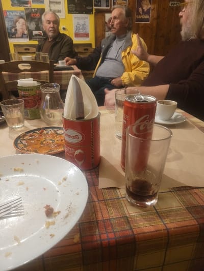 The image depicts three older adult males seated around a plaid-covered table in a casual eatery or tavern in Κωστήτσι, Greece. In the immediate foreground, a white ceramic plate with food residue and a silver fork is positioned next to a clear glass with a small amount of liquid. A decorative ceramic plate containing an orange-red dish is visible behind the white plate. A red San Marzano branded tomato can, repurposed as a napkin holder, stands upright on the table, containing white paper napkins and a dark, cylindrical object. To the left of the napkin holder, a clear glass pitcher is present. On the right side of the table, a red Coca-Cola can, an empty clear drinking glass, and a white coffee cup with a dark liquid residue on a saucer are visible.
The man on the far left has grey hair and a beard, wearing a brown jacket, and is looking towards the right side of the frame. The man in the middle has a mustache and is wearing a yellow jacket over a darker garment, also looking right. The man on the far right, partially visible, has long grey hair and a beard, wearing a dark reddish-brown long-sleeved shirt, with his right hand raised in an open-palm gesture, suggesting active communication. The background features light yellow walls partially covered with dark wood paneling, adorned with various posters and signs, including text such as "Ο ΓΑΡΟΣ", "ZOO", and "WC".